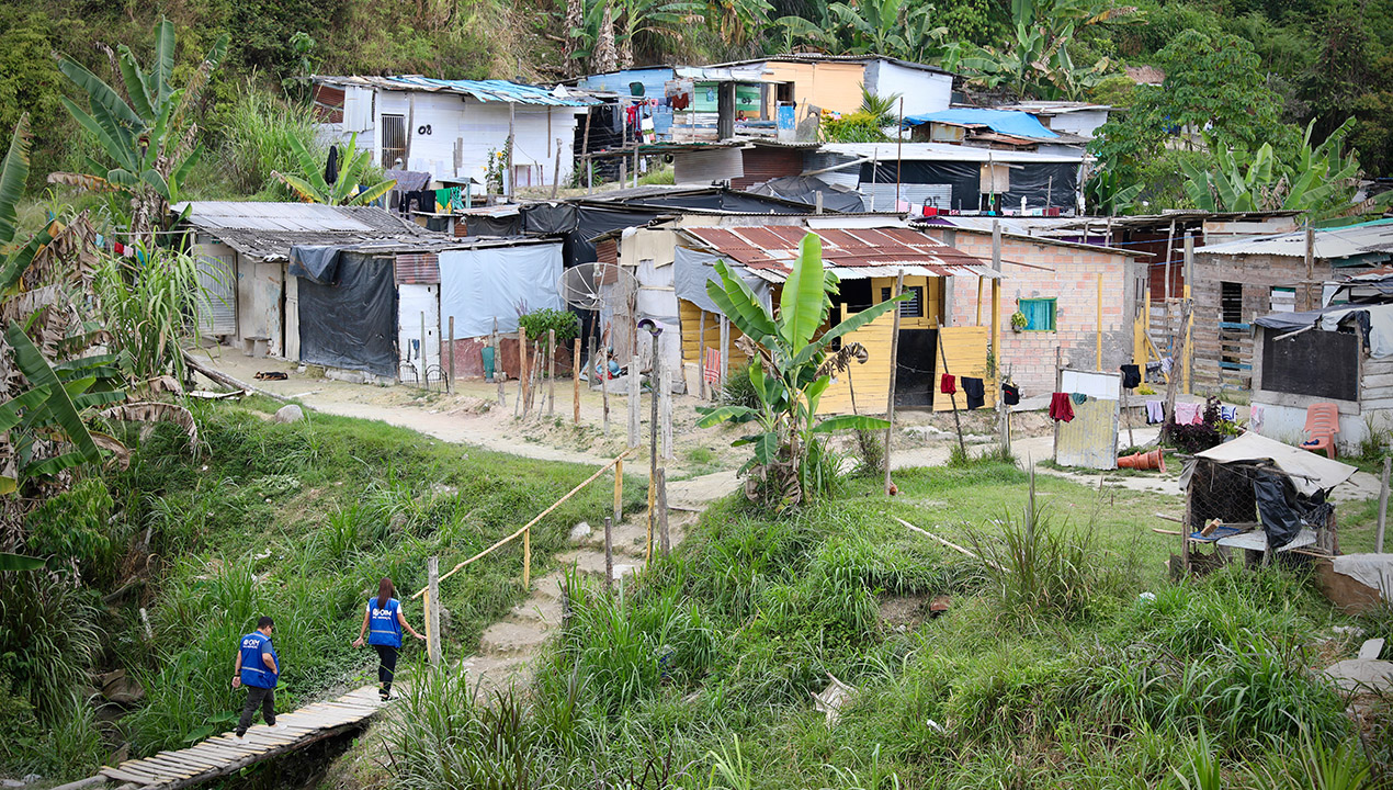 Vista aérea da Vila Esperança: um conjunto de casas em uma ocupação espontânea, sendo acessada por duas pessoas com colete da OIM, passando por uma ponte improvisada